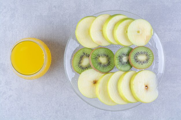 Rodajas de manzana y kiwi sobre un pedestal de vidrio junto a un vaso de jugosa naranja, sobre la mesa de mármol.