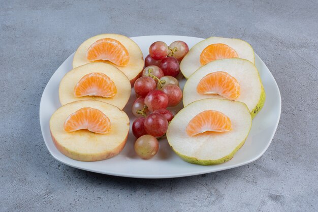 Rodajas de manzanas, peras, mandarinas y uvas en un plato sobre fondo de mármol.