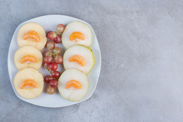 Rodajas de manzanas, peras, mandarinas y uvas en un plato sobre fondo de mármol.