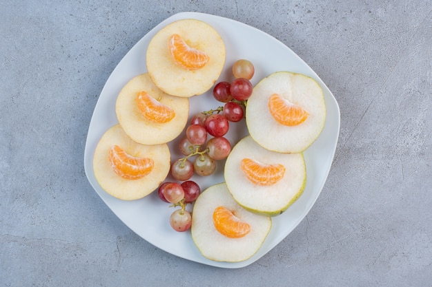 Rodajas de manzanas, peras, mandarinas y uvas en un plato sobre fondo de mármol.