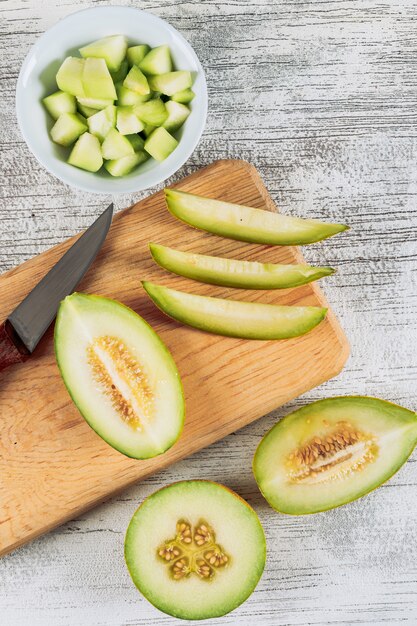 Rodajas de melón en una tabla de cortar de madera con melón en un tazón y un cuchillo plano sobre un fondo de piedra blanca