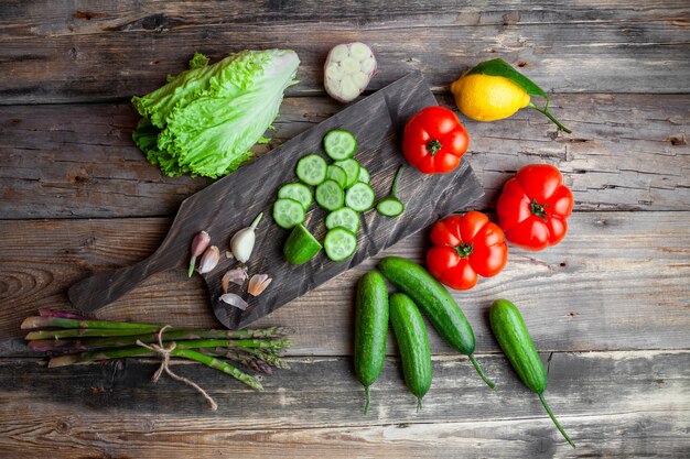 Rodajas de pepino en una tabla de cortar con tomates, lechuga, limón vista superior sobre un fondo oscuro de madera