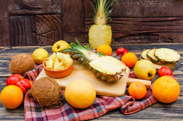 Rodajas de piñas jugosas con cocos, duraznos, membrillos y cítricos en tablas de madera y un tazón en la superficie de madera del grunge, tela de picnic y baldosas de piedra, endecha plana.