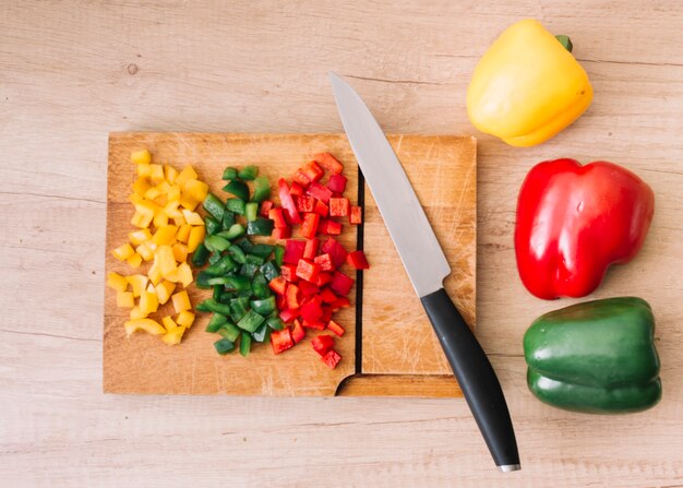 Rojo entero y picado; verde; Pimientos amarillos en tabla de cortar con cuchillo afilado contra el telón de fondo de madera