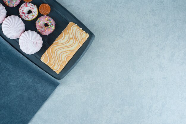 Rollo de pastel, galletas, donas y mermeladas sobre un tablero azul marino sobre mármol.