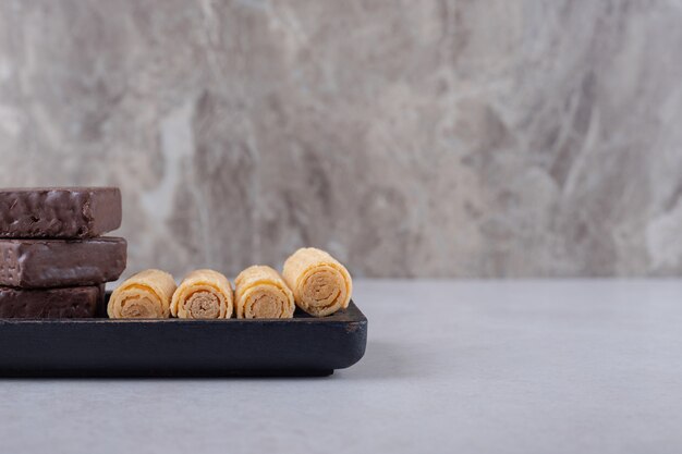Rollos de obleas deliciosas y obleas recubiertas de chocolate en una placa de madera sobre el mármol.