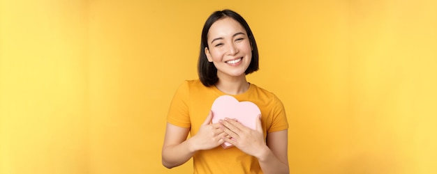 Foto gratuita romance y día de san valentín feliz hermosa mujer asiática sosteniendo una gran tarjeta de corazón y sonriendo de pie o...