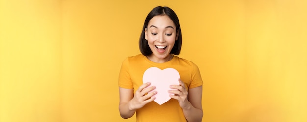 Romance y día de san valentín feliz hermosa mujer asiática sosteniendo una gran tarjeta de corazón y sonriendo de pie o