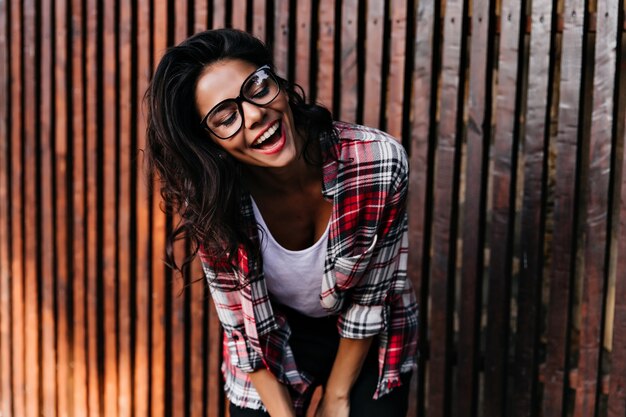 Romántica chica bronceada riendo mientras posa junto a la valla de madera. Señora feliz de pelo oscuro en camisa a cuadros sonriendo con los ojos cerrados.