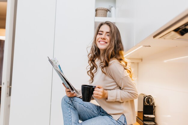 Romántica chica morena en jeans sosteniendo taza negra y sonriendo