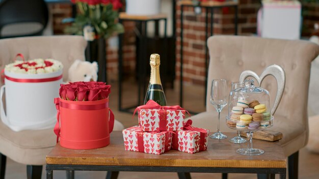 Romántica configuración de lujo para el día de San Valentín con cajas de regalo, champán y macarons en una mesa de café con un elegante ramo de rosas. Sala de estar con flores de decoración temática de amor sorpresa y oso de peluche.