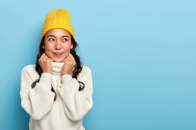 Romántica hermosa adolescente asiática recuerda un momento agradable, usa un sombrero amarillo y un suéter blanco cálido, mantiene las manos en el cuello, está sumido en pensamientos durante el frío día de invierno