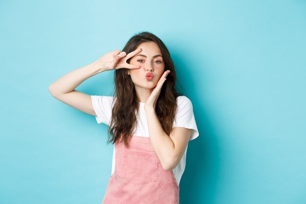 Romántica y hermosa joven enviando beso al aire, frunciendo los labios y mostrando un gesto de paz con signo de v cerca del ojo, de pie sobre fondo azul.