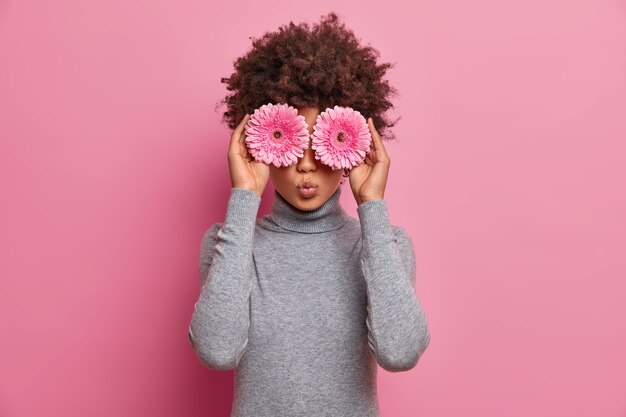 Romántica hermosa mujer de pelo rizado mantiene gerberas rosadas sobre los ojos, tiene un estado de ánimo primaveral, vestida con un cuello de tortuga gris informal, va a hacer un ramo de flores