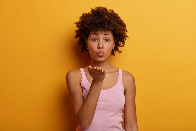 Romántica joven étnica rizada mantiene los labios doblados, envía besos al aire, expresa amor y coquetea con su novio, mira con ternura, posa contra la pared amarilla. Concepto de lenguaje corporal