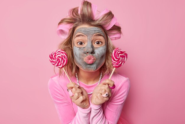 Romántica joven europea mantiene los labios redondeados envía mwah sostiene dos piruletas cerca de la cara aplica rodillos para el cabello y máscara de belleza para eliminar las líneas finas usa un vestido aislado sobre fondo rosa.