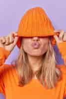 Foto gratuita romántica joven mujer irreconocible mantiene los labios redondeados quiere besarte cubre los ojos con sombrero de punto sopla mwah vestida con poses de puente naranja sobre fondo púrpura expresiones faciales