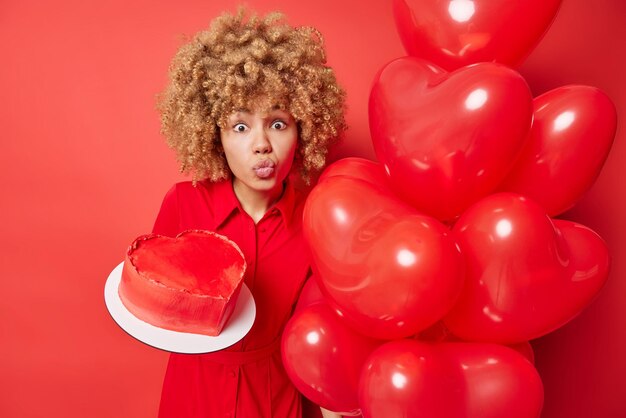 Romántica mujer de cabello rizado mantiene los labios plegados sostiene un delicioso pastel en forma de corazón y globos inflados viste un vestido aislado sobre un fondo rojo vívido Concepto de celebración de vacaciones de personas