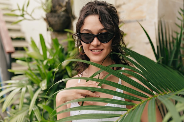 Romántica mujer feliz con cabello corto y rubio ojos cerrados y disfrutando de las vacaciones en un día caluroso de verano en la isla en la pared de plantas exóticas