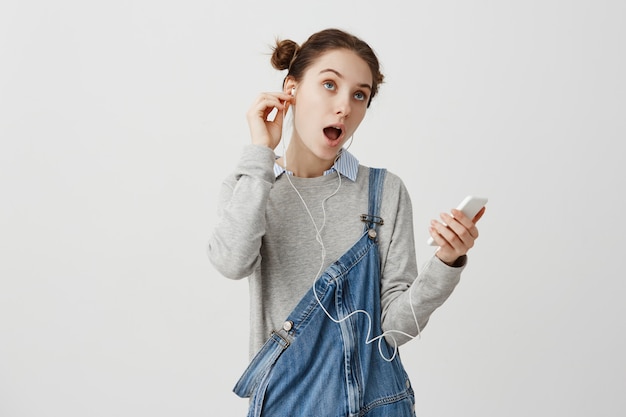 Romántica mujer joven con el pelo en doble bollos cantando mientras disfruta de la música de su teléfono celular. Amante de la música femenina descansando después de un día duro escuchando canciones con auriculares. Concepto de hobby