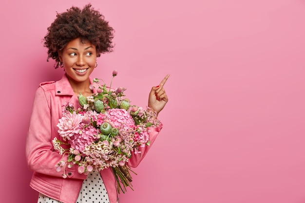 Romántica mujer joven positiva con cabello afro apunta el dedo índice a un lado, tiene un bonito ramo de flores mixtas