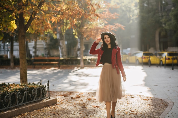 Romántica mujer de pelo negro en falda larga y exuberante disfrutando del sol en el parque de otoño