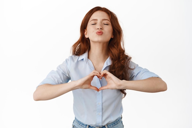 Romántica novia pelirroja inclinándose por un beso, mostrando el corazón Te amo firmar, siendo sincera y linda, de pie contra el fondo blanco