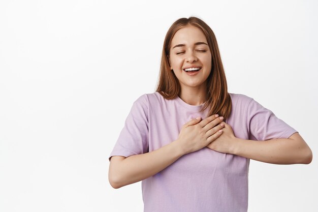 Romántica y soñadora joven rubia, mirada apasionada, cerrar los ojos y tomarse de las manos en el corazón, soñar despierto, imaginar una pared blanca y hermosa.