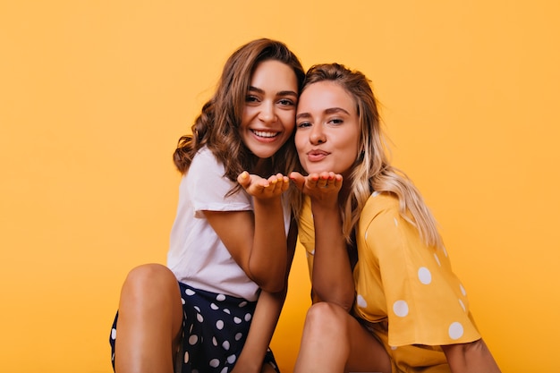 Románticas chicas bronceadas enviando besos al aire. Alegre amigas expresando amor con una sonrisa.