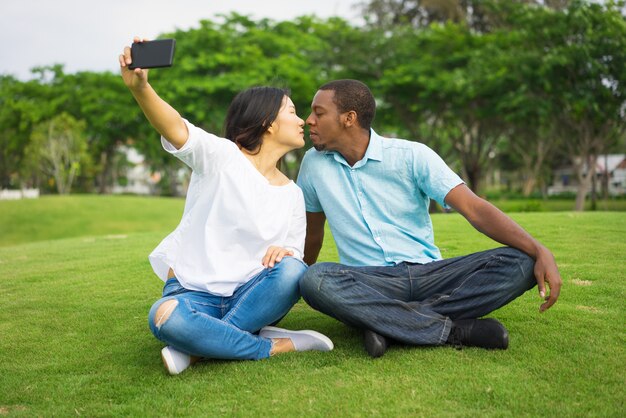 Romántico joven pareja besándose y tomando selfie con teléfono móvil.