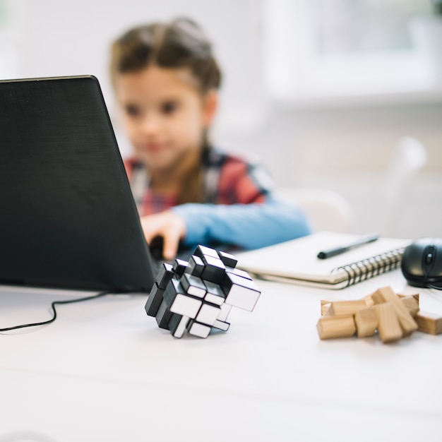 Foto gratuita rompecabezas de cubo frente a una niña usando una computadora portátil en el escritorio blanco