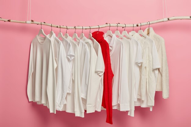Ropa casual blanca para mujer dispuesta en perchas, suéter cálido de punto rojo se destaca de toda la colección. Conjunto de vestuario colgando sobre fondo rosa. Armario de casa. Estilo clásico. Tienda de moda