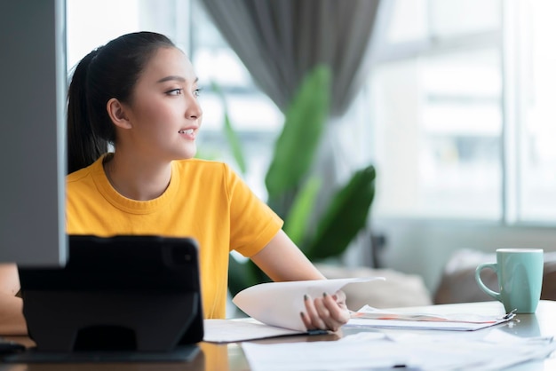Ropa casual femenina asiática activa que trabaja en casa en cuarentena trabajo remoto en línea siéntese y relájese en el escritorio de trabajo con un fondo interior alegre y almeja