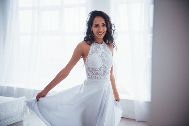 Ropa clásica para baile y fiestas de lujo. Hermosa mujer vestida de blanco se encuentra en la sala blanca con luz del día a través de las ventanas