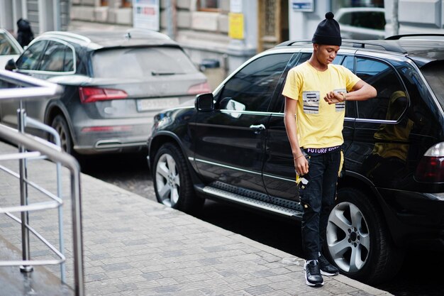 Ropa de hombre africano con sombrero negro posado al aire libre contra el coche de negocios