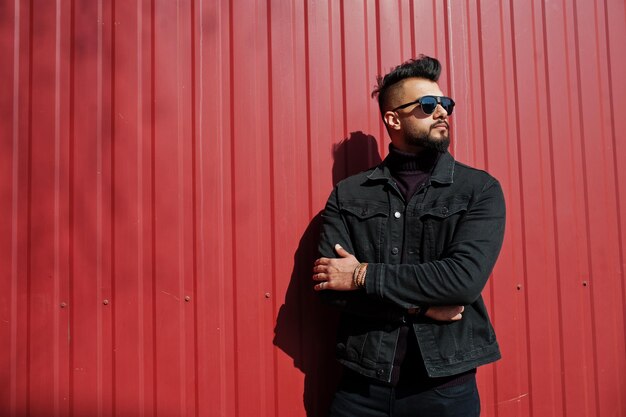 Ropa de hombre árabe de moda con chaqueta de jeans negros y gafas de sol posadas contra el fondo de la pared de acero rojo Chico modelo árabe elegante y de moda