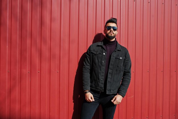 Ropa de hombre árabe de moda con chaqueta de jeans negros y gafas de sol posadas contra el fondo de la pared de acero rojo Chico modelo árabe elegante y de moda