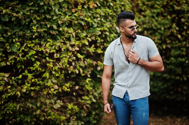 Ropa de hombre de barba árabe alto de moda en jeans de camisa y gafas de sol posadas en el parque contra la vegetación