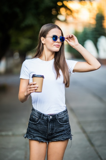 Ropa de mujer joven en definitiva, camisa blanca y gafas de sol caminando con café para salir a la calle