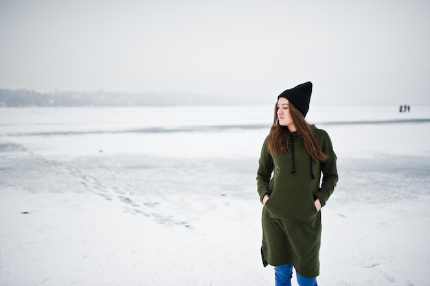 Ropa de niña divertida en pantalones vaqueros largos de sudadera verde y sombreros negros en el lago congelado en el día de invierno