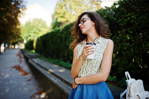 Ropa de niña con estilo rizado en blusa de falda de jeans azules y gafas posadas en la calle de la ciudad con una taza de café