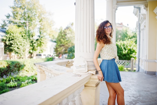 Ropa de niña con estilo rizado en blusa de falda de jeans azules y gafas posadas cerca de la antigua casa vintage