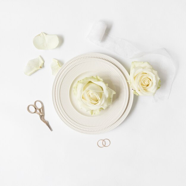 Rosa fresca en un plato con cinta; tijera y anillos de boda sobre fondo blanco