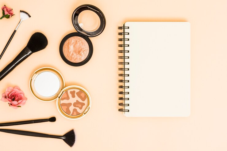 Foto gratuito rosa; pinceles de maquillaje y polvos compactos cerca de la libreta espiral sobre fondo beige