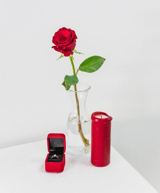 Rosa roja fresca en florero y caja actual con anillo cerca de vela en mesa