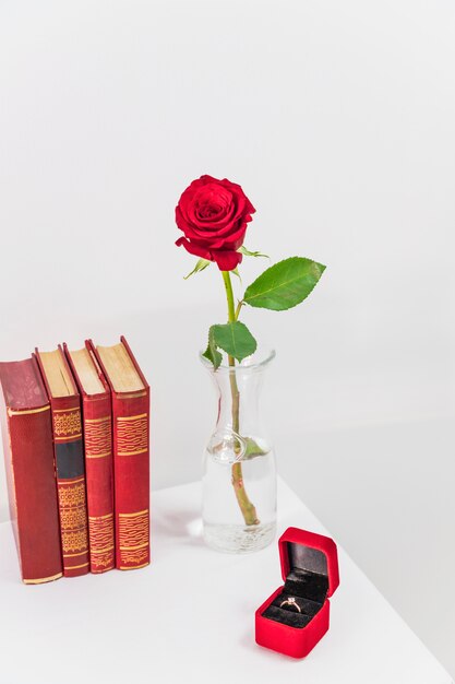 Rosa roja fresca en el florero cerca de la caja actual con anillo y libros en la mesa