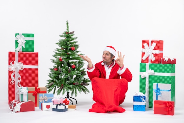 Rostro agrio joven vestido como Papá Noel con regalos y árbol de Navidad decorado sentado en el suelo haciendo gesto de anteojos apuntando algo sobre fondo blanco