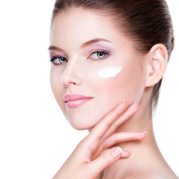 Rostro de belleza de mujer joven con crema cosmética en la mejilla. Concepto de cuidado de la piel. Retrato de detalle aislado en blanco.