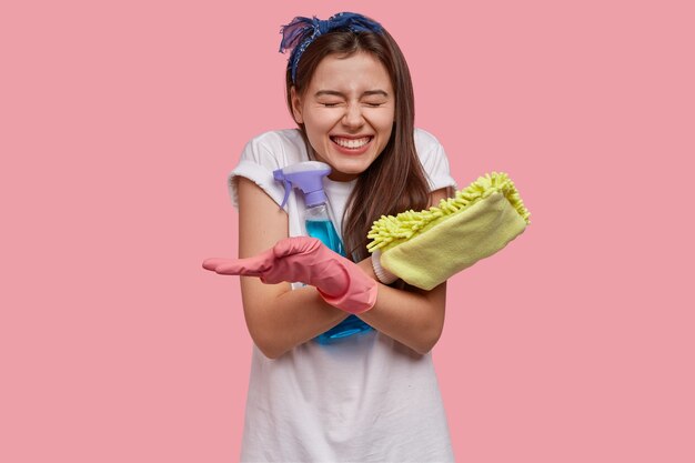 El rostro femenino positivo sonríe, tiene una amplia sonrisa, dientes blancos perfectos, lleva un trapo con un agente de limpieza, está de buen humor