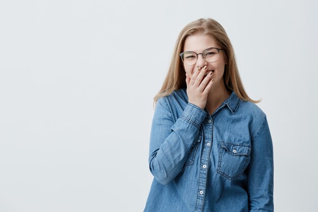 Rostro humano expresiones y emociones. Joven rubia positiva y encantadora riéndose sinceramente de una broma divertida, mirando a la cámara, vestida con una camisa vaquera y gafas, ocultando la cara detrás de la palma